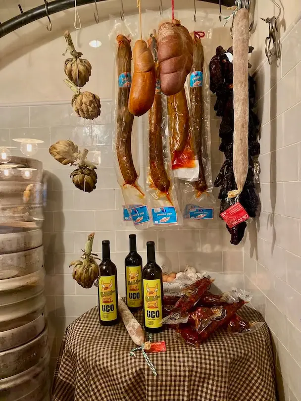 Various sausages are hanging from the ceiling in this room next to the artichokes on the left. Olive oil in bottles can be found on the table.