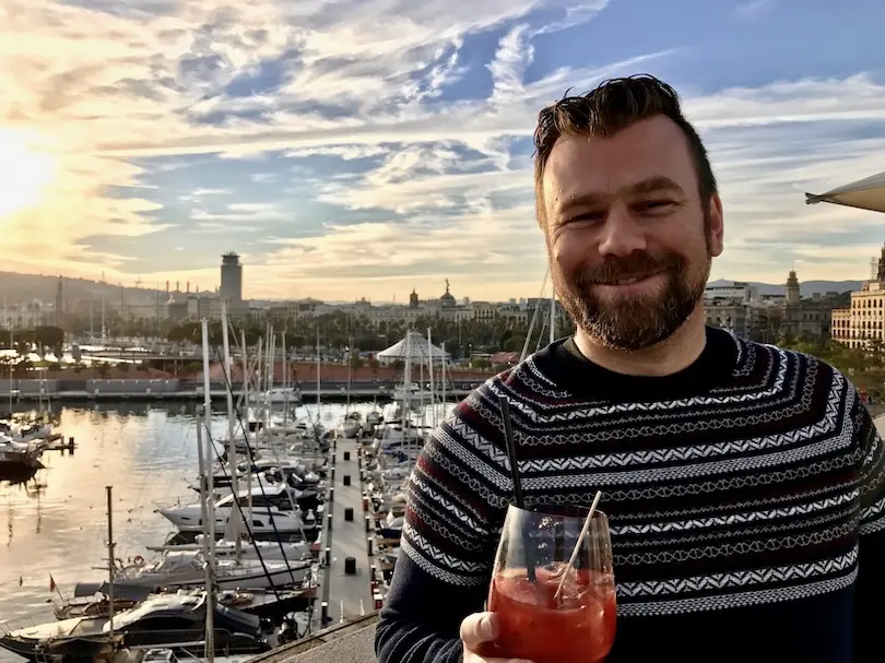 Tour guide Christian with a drink in his hand is standing on a roof top terrace with his back towards the old town of Barcelona.