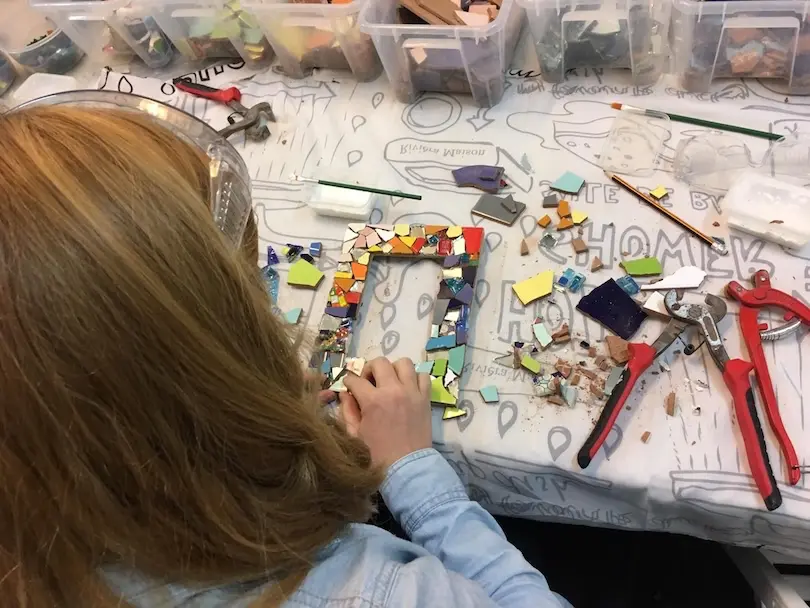 A lady is making a picture frame out of colorful broken mosaic tiles. Next to her she has her tools.
