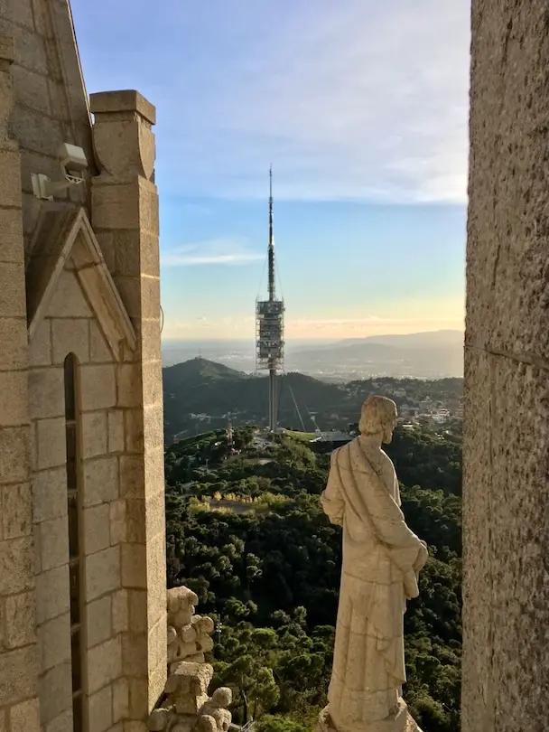 Barcelona´s telecommunication tower can be seen in the distance. A big statue of a saint made out of stone is overlooking the panoramic view of the Collserola natural park.