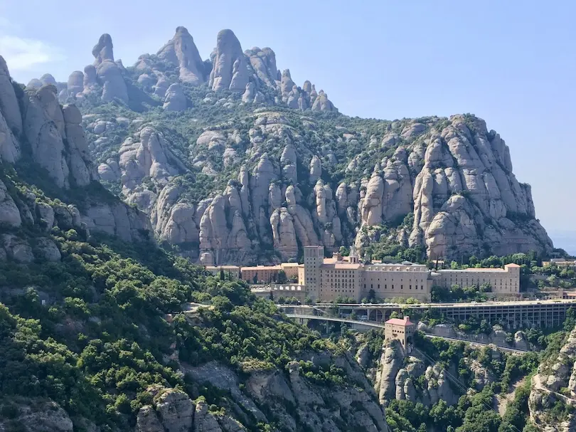 The monastery of Montserrat is located beneath high rock formations under a blue sky.